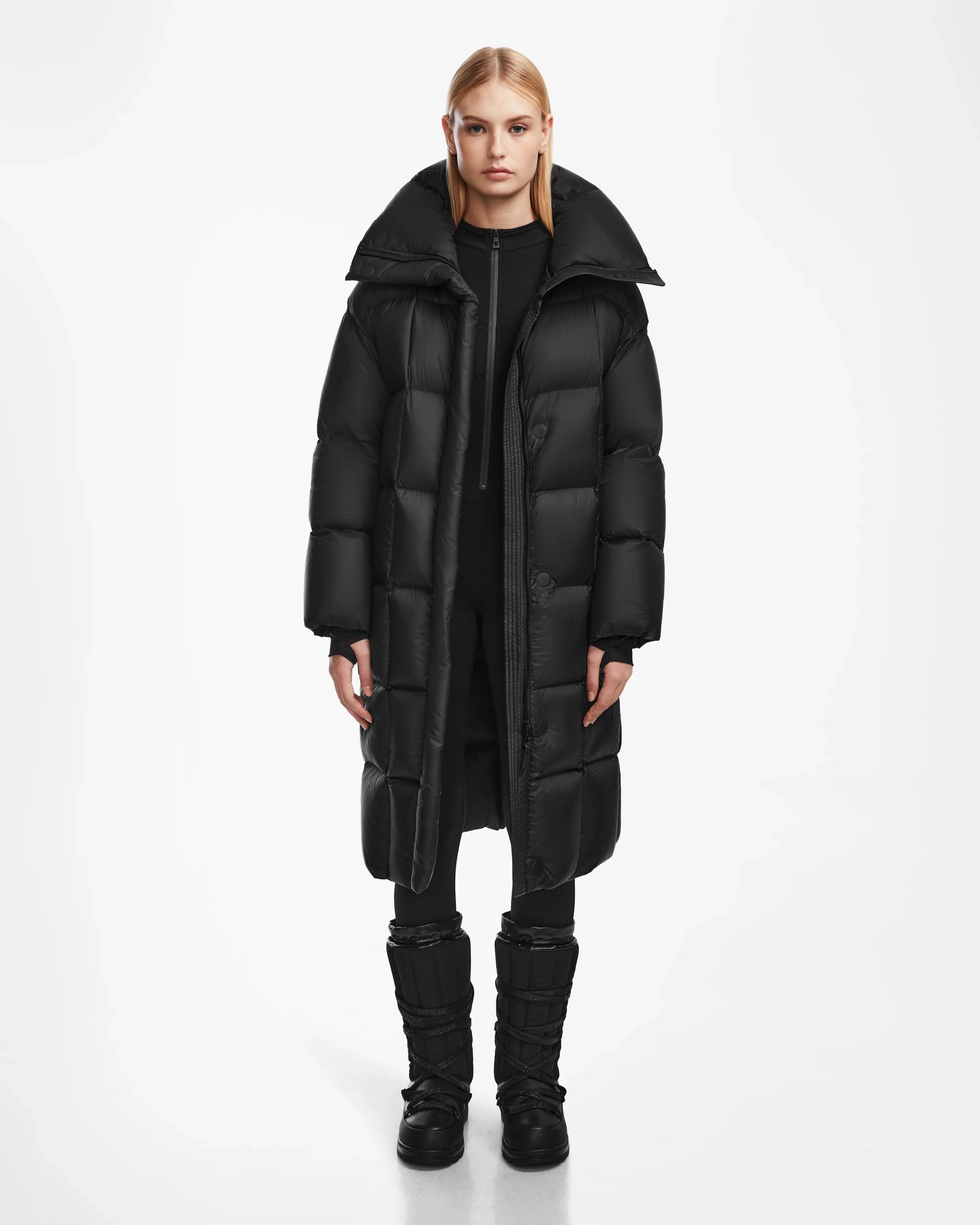 A person faces forward against a plain white background, wearing the Rudsak SASH - 8124536 Black womens long down puffer coat, paired with black pants and black boots.