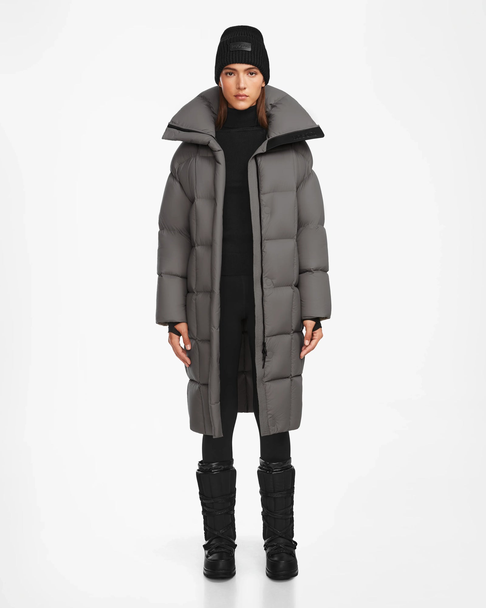 A woman models the Rudsak SASH - 8124536 Light Grey womens long down puffer coat, styled with a black beanie, pants, and winter boots. She stands against a plain white background, facing forward with a neutral expression.