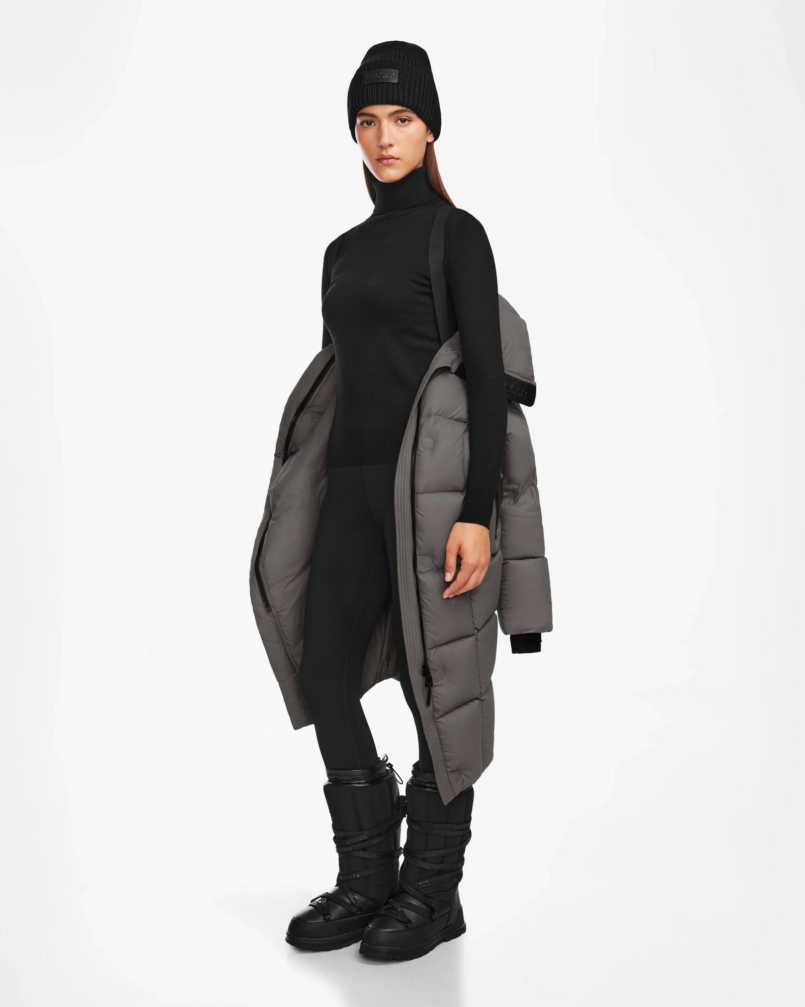 A woman stands facing forward against a plain white background, wearing a black beanie, turtleneck, leggings, winter boots, and the Rudsak SASH - 8124536 Light Grey womens long down puffer coat draped over her shoulders.