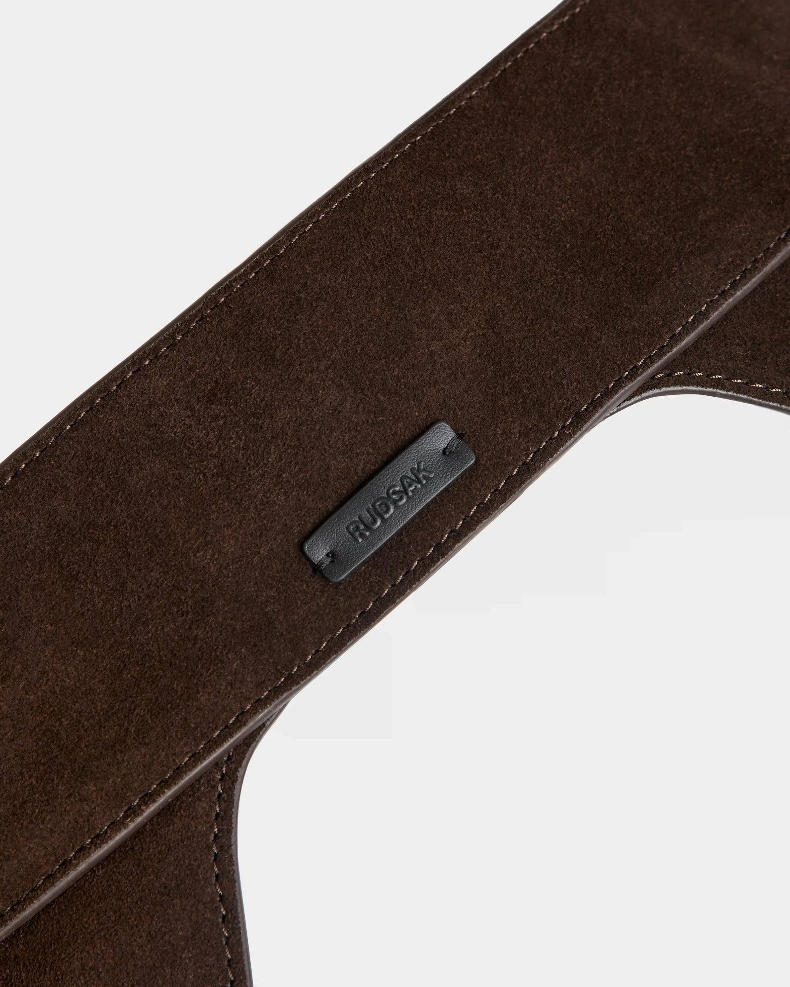 Close-up of the SIMONA S - 8424523 Espresso womens suede waist belt by Rudsak, featuring visible stitching and a small black tag with the brand name in raised letters. The background is plain and light-colored.