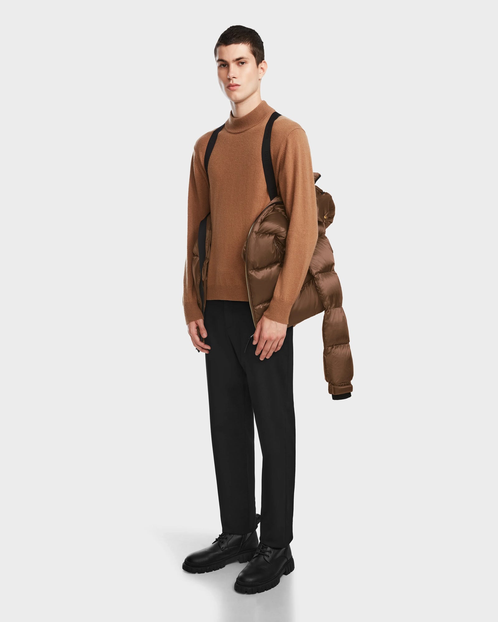 A young man faces forward in black pants and shoes, with a brown sweater and the Rudsak VINCE S - 6124560 Antique Bronze mens quilted down puffer jacket draped over one shoulder, standing against a plain white background.