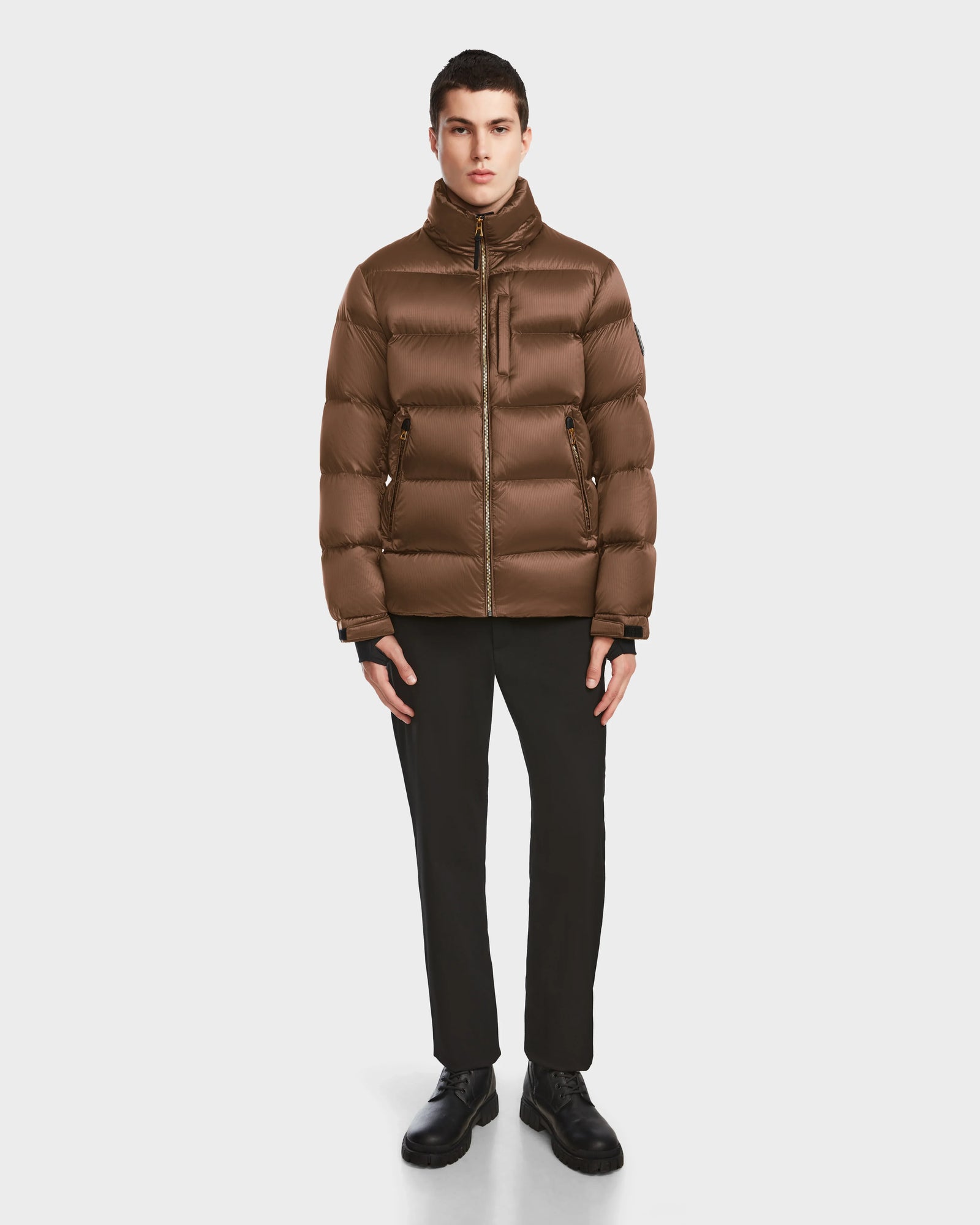 A person faces forward wearing the Rudsak VINCE S - 6124560 Antique Bronze mens quilted down puffer, paired with black pants and shoes, against a plain white background.