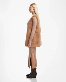 A woman with long blonde hair stands in profile wearing the Rudsak YOLANDA - 8124790 Camel cable-knit dress, styled with a vegan leather belt, faux fur vest, and brown ankle boots—an ideal look for fall and winter.