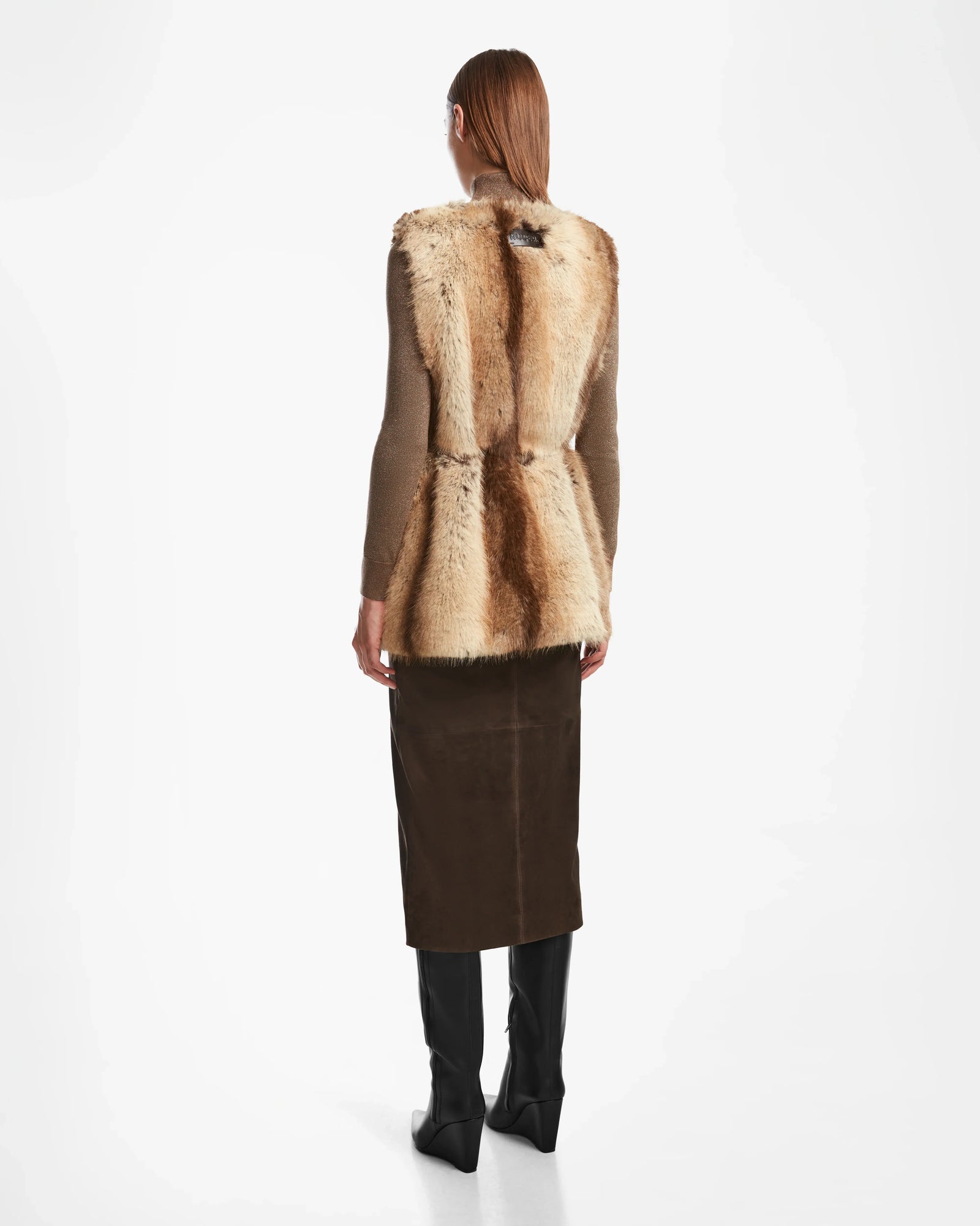 A woman faces away wearing the Rudsak YOLANDA - 8124790 Mocha womens faux fur vest over a brown top, styled with a vegan leather belt, dark brown skirt, and black knee-high boots against a white background.