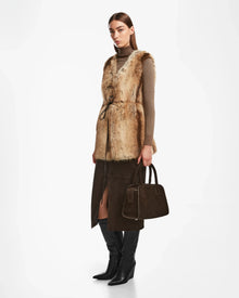 A woman wears the Rudsak YOLANDA - 8124790 Mocha faux fur vest over a tan turtleneck, dark brown slit skirt, black knee-high boots, and holds a matching handbag—an ideal addition to your fall-winter wardrobe.