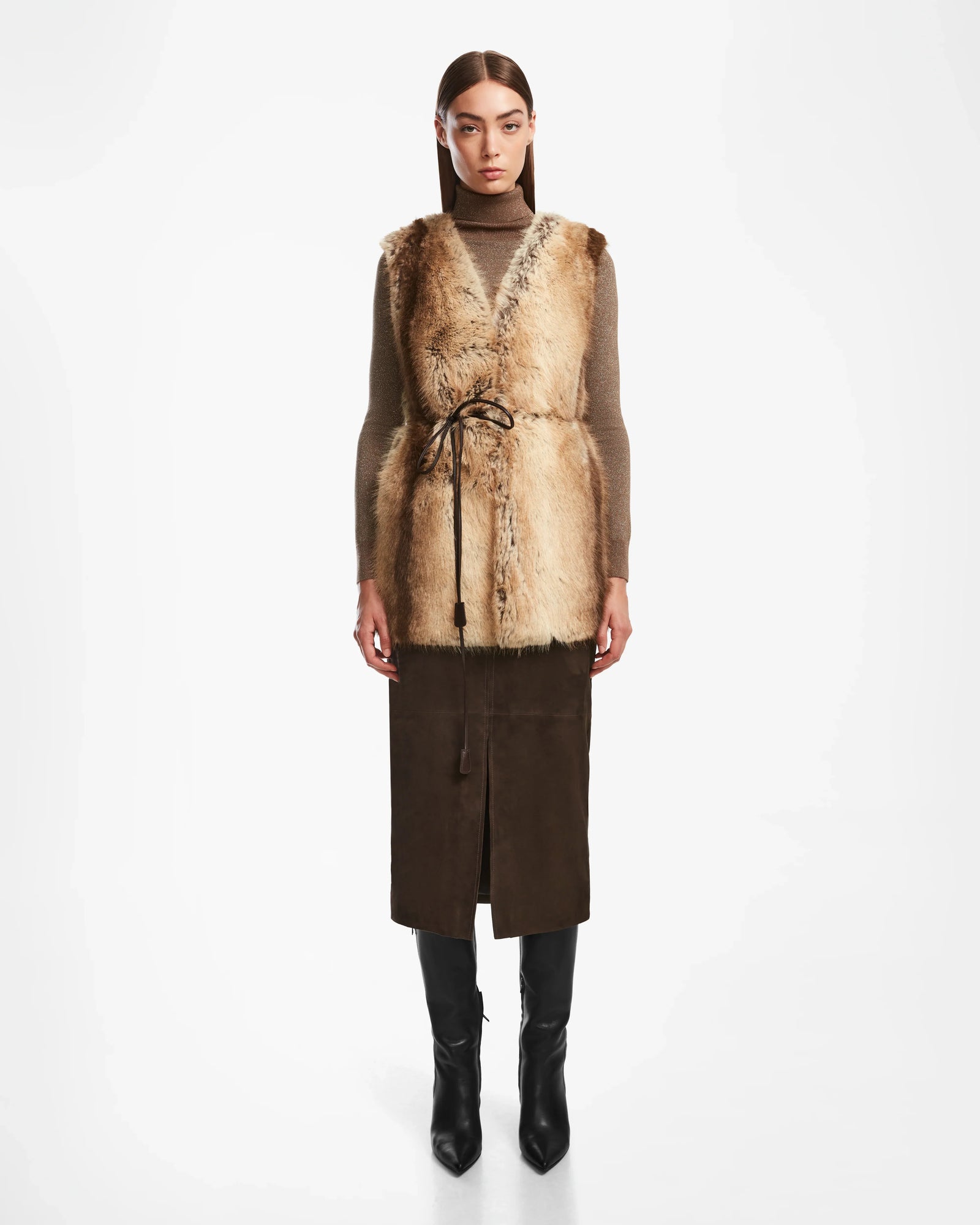 A woman faces forward wearing the Rudsak YOLANDA - 8124790 Mocha faux fur vest over a brown turtleneck, a dark brown suede skirt with a vegan leather belt, and black knee-high boots against a plain white background.