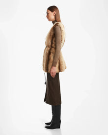 A woman stands in profile against a white background, wearing the YOLANDA - 8124790 Mocha faux fur vest by Rudsak over a brown long-sleeve top, dark brown midi skirt with vegan leather belt, and black knee-high boots.