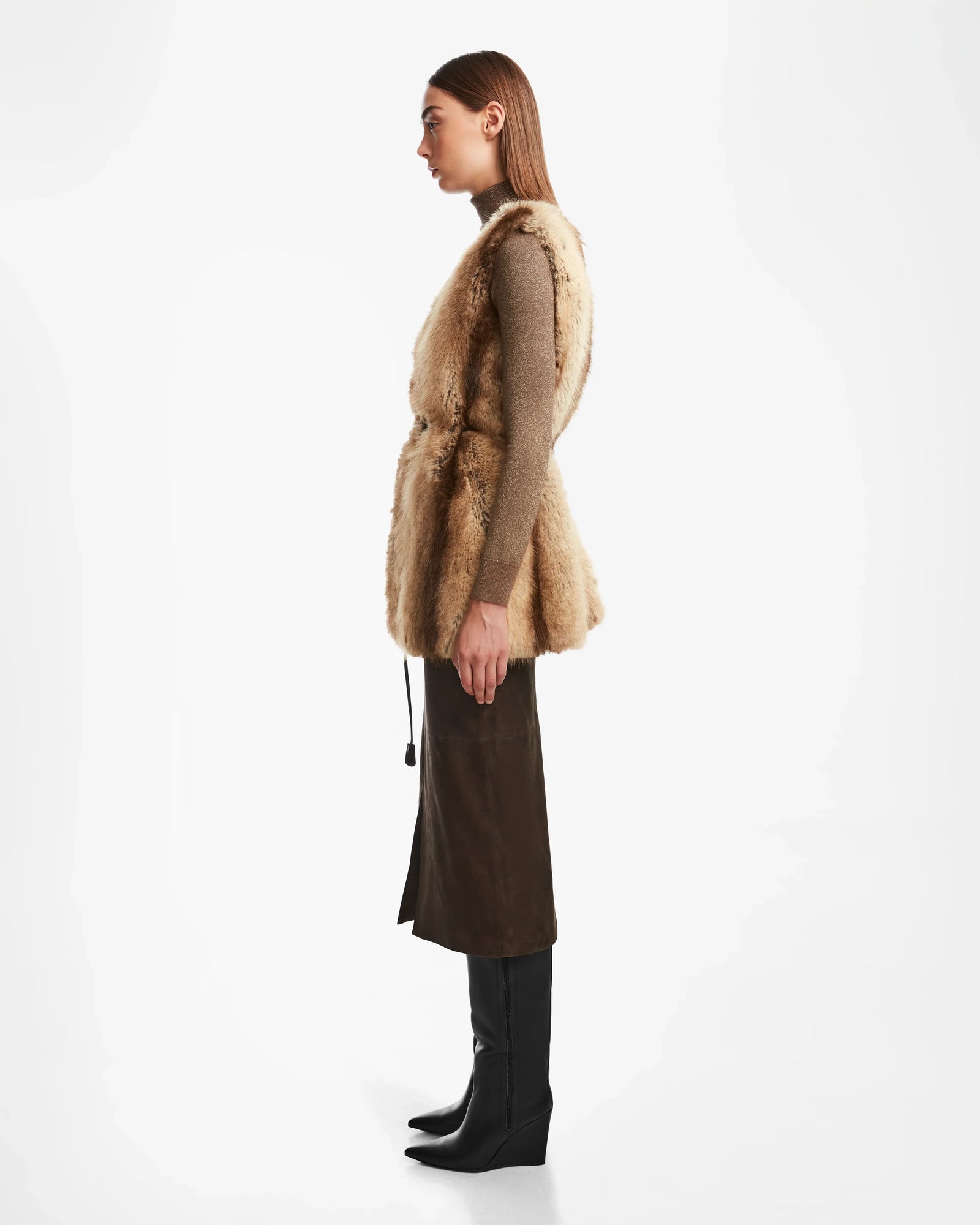 A woman stands in profile against a white background, wearing the YOLANDA - 8124790 Mocha faux fur vest by Rudsak over a brown long-sleeve top, dark brown midi skirt with vegan leather belt, and black knee-high boots.