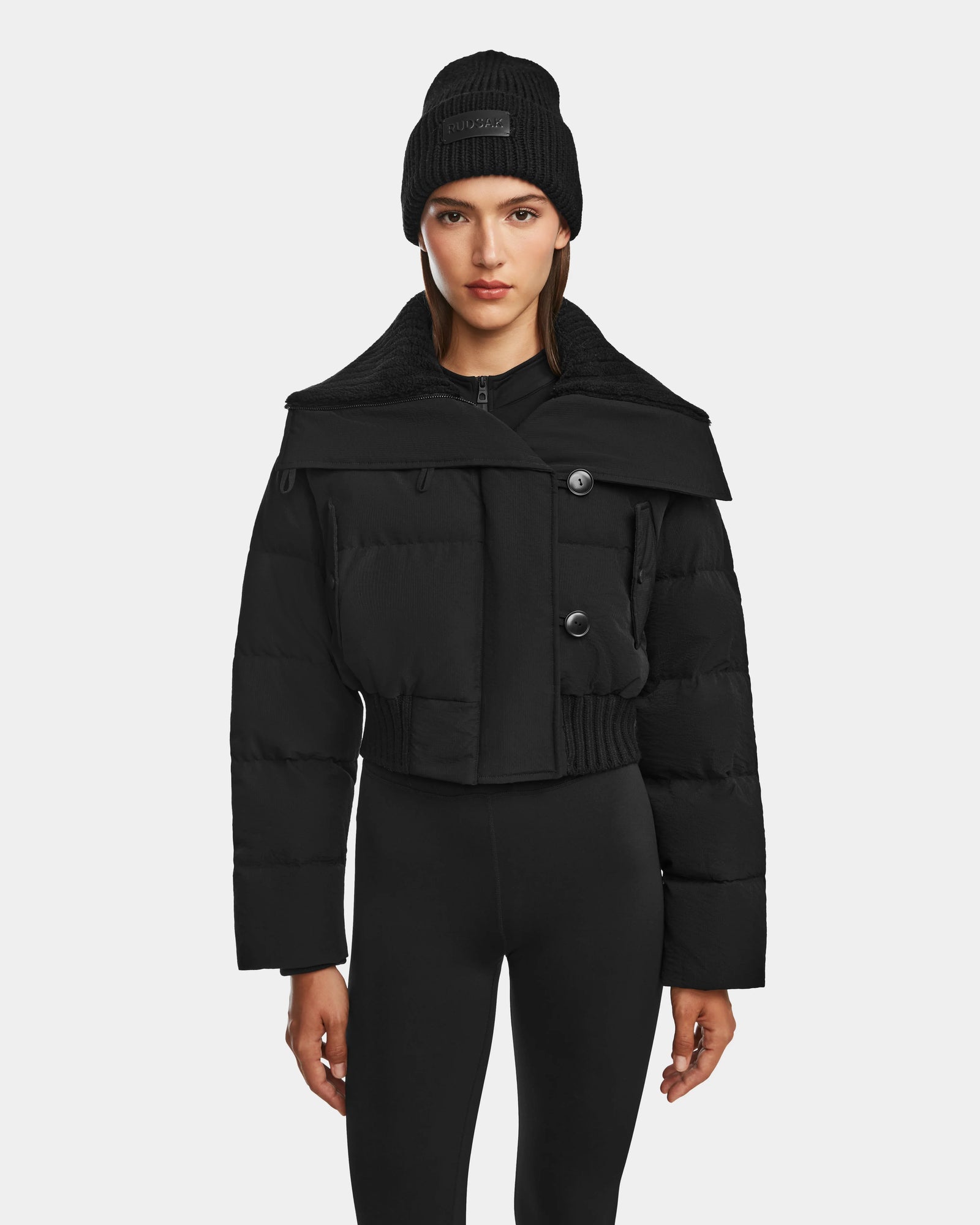 A woman models Rudsaks YVETTE - 8124710 Black women’s crop down puffer jacket with large buttons, paired with a black beanie and leggings, standing against a plain white background.