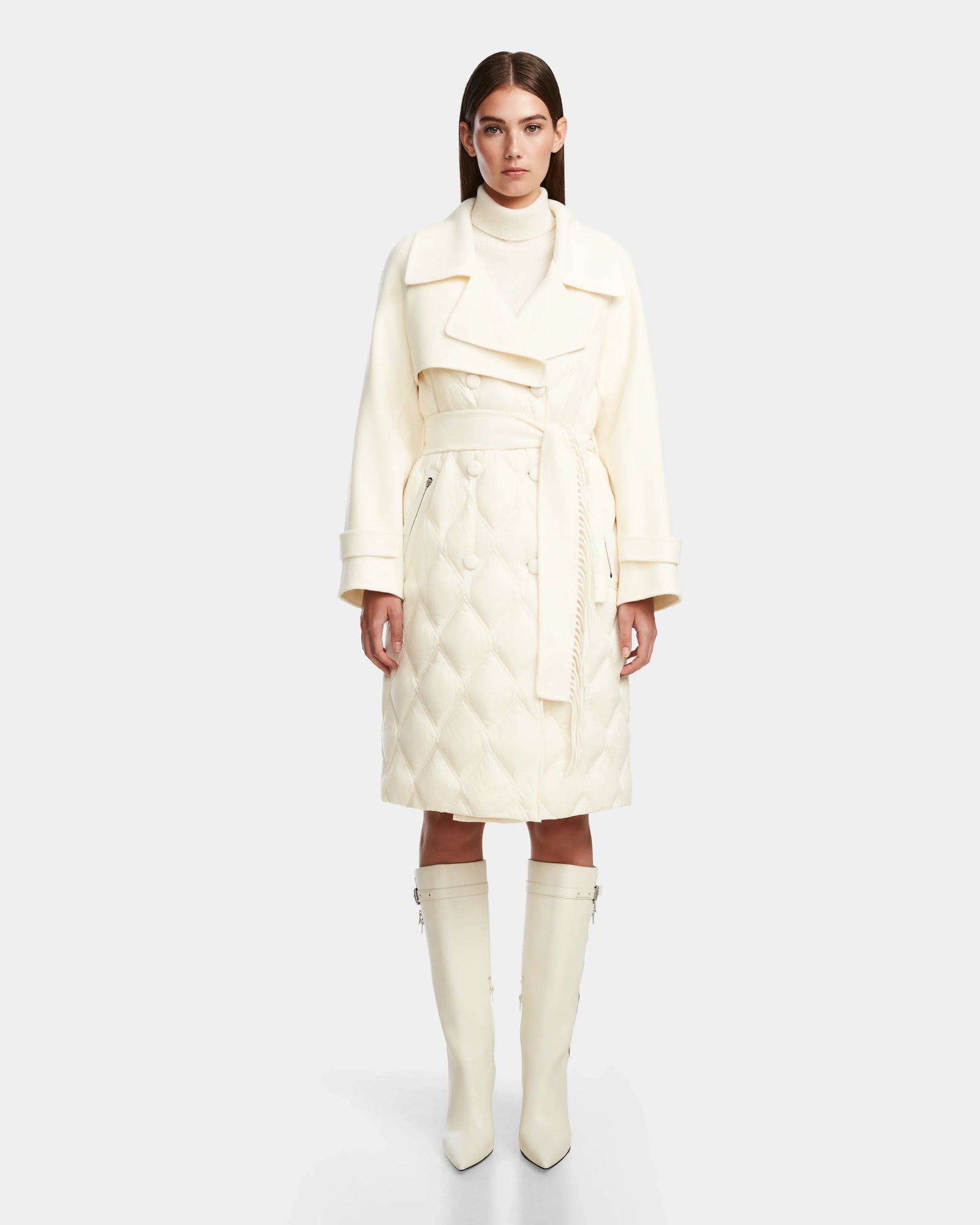 A woman with straight brown hair and a neutral expression stands facing forward, wearing the Rudsak ZOE - 8124711 Iceland cream quilted down trench with a wide collar, belted waist, and matching knee-high boots.
