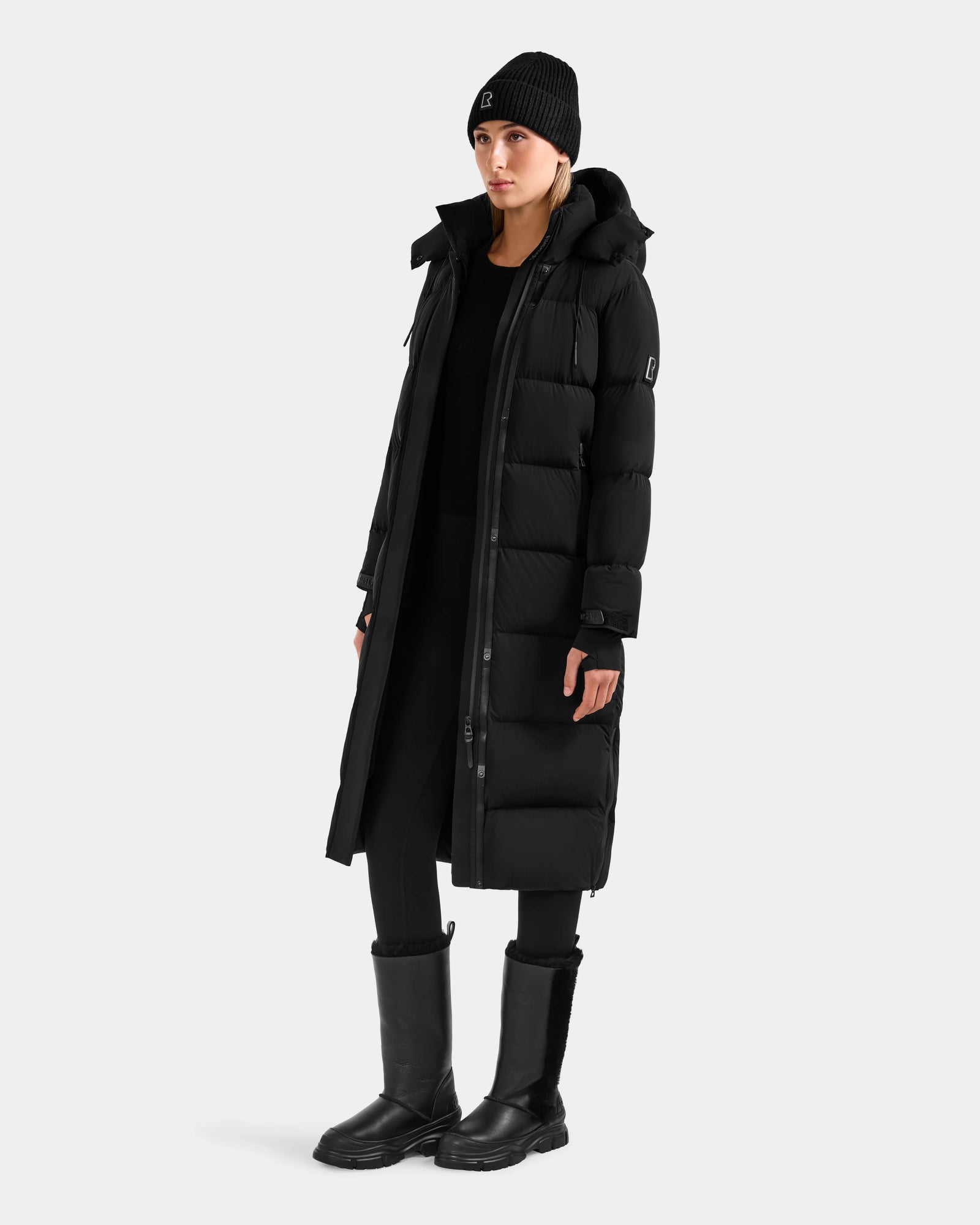 Wearing the Rudsak ALESSANDRA L - L125553 Black quilted down puffer coat, paired with a black beanie, leggings, and boots, this look against a light gray background is the ultimate winter essential.