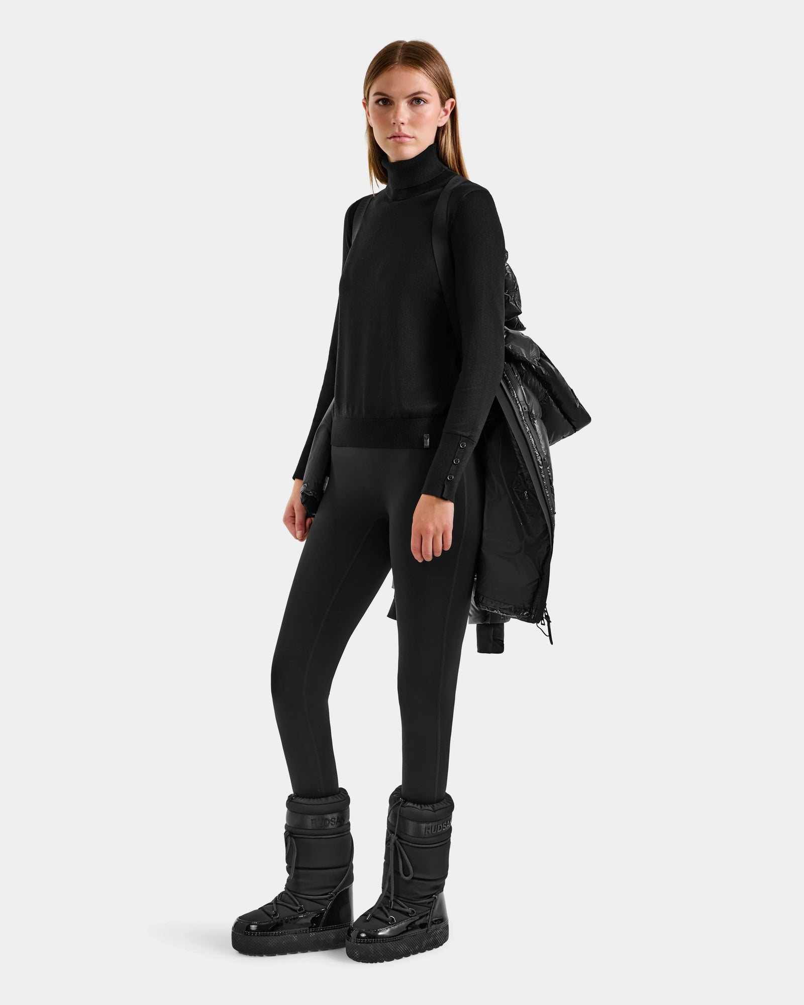 A woman stands against a neutral background wearing the Rudsak ALESSANDRA S-S - L125560 Black turtleneck, black leggings, black winter boots, and holding a glossy puffer jacket over her shoulder.