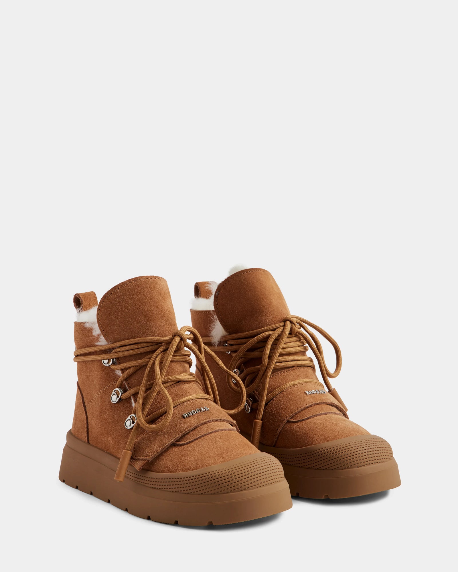 The Rudsak ALEXEY - L225025 Tan women’s suede winter boots feature thick rubber soles, metal eyelets, tan laces, and white fur trim at the top. Shown side by side on a light gray background.