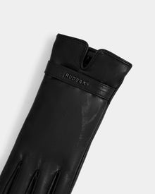 A close-up of the Rudsak ALEXIA - L425543 Black women’s leather glove with visible stitching and an embossed brand on the wrist band, laid flat on a light gray background—perfect for cold-weather wear.