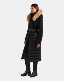 A woman wears the Rudsak ALISA L - L125574 Black long quilted puffer coat with RDS-certified down, belted waist, and tan faux fur-trimmed hood, paired with black pants and ankle boots against a plain light gray background.