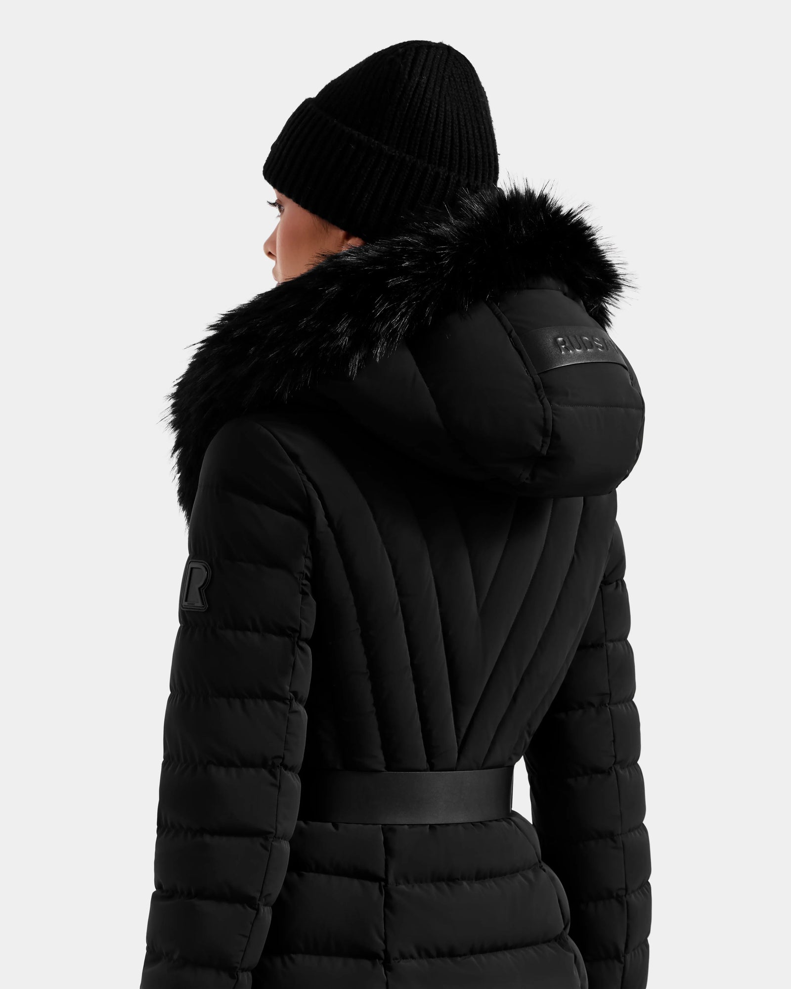 A person in the Rudsak ALISA M - L125586 Black quilted down parka with faux fur-lined hood and a black knit beanie, shown from the back against a plain light background.