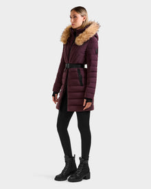 A woman wears the Rudsak ALISA M - L125586 Bordeaux quilted down parka with vegan leather belt and faux fur-lined hood, paired with black leggings and lace-up boots, standing against a plain light background.