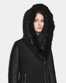 A woman models the Rudsak AURELLE - L124538 Black down parka, featuring a large faux fur hood and vegan leather sleeves, as she looks at the camera against a plain light background.