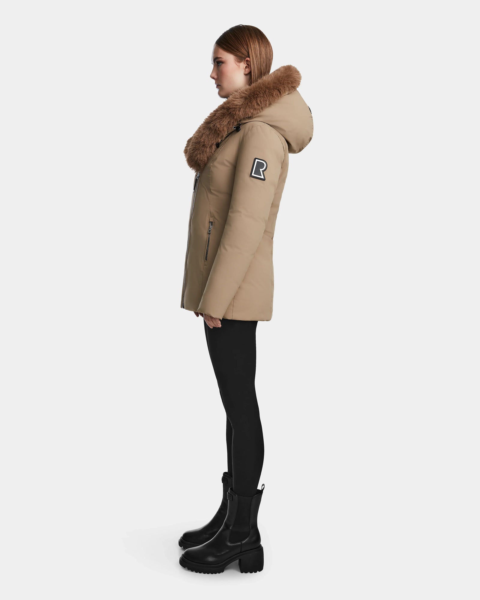 A woman stands in profile against a plain white background, wearing the Rudsak AURORE - L123561 Sand winter parka with a large fur-lined hood, black leggings, and boots. The fitted coat features an "R" patch on the upper sleeve.