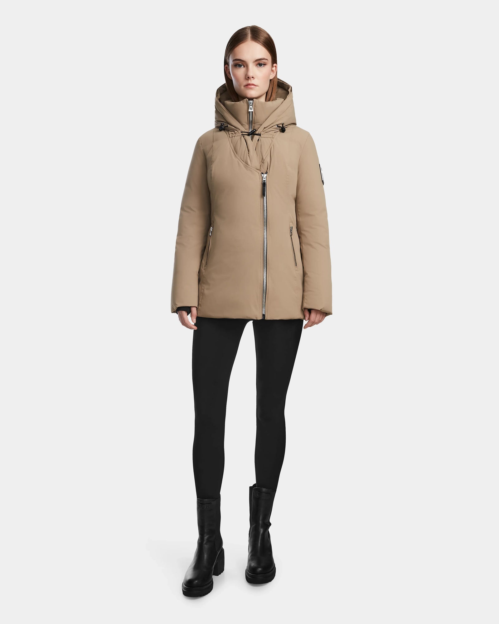 A woman stands against a plain background wearing the Rudsak AURORE - L123561 Sand parka, paired with black leggings and ankle boots. She faces forward with a neutral expression, her hands at her sides.