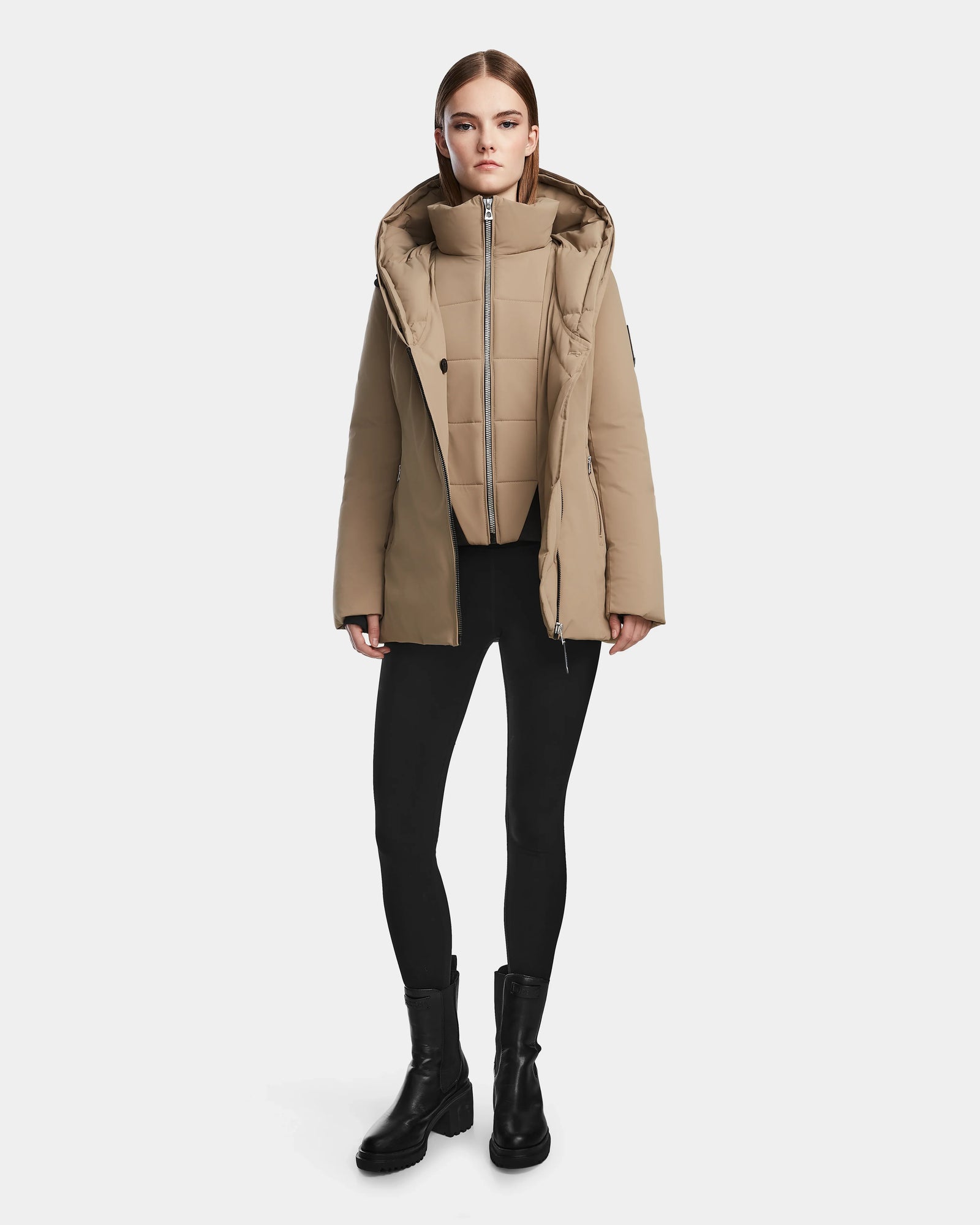 A woman faces forward against a white background, wearing the Rudsak AURORE - L123561 Sand weather-resistant parka with a hood, paired with black leggings and black ankle boots.