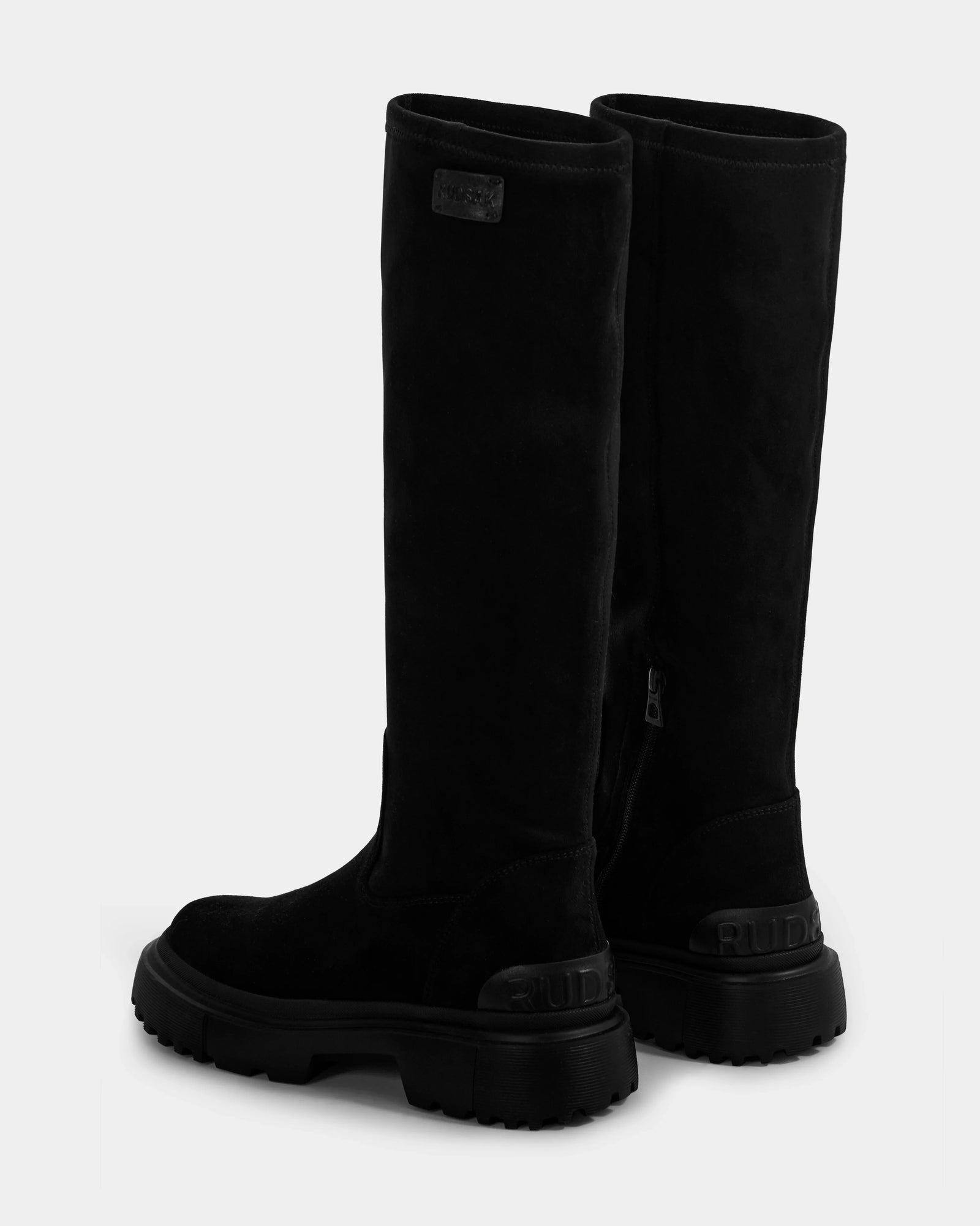 The AVOLA - L225514 Black boots by Rudsak are tall women’s knee-high boots with thick, ridged soles and the brand name embossed on the heels, displayed facing slightly away on a plain light background.