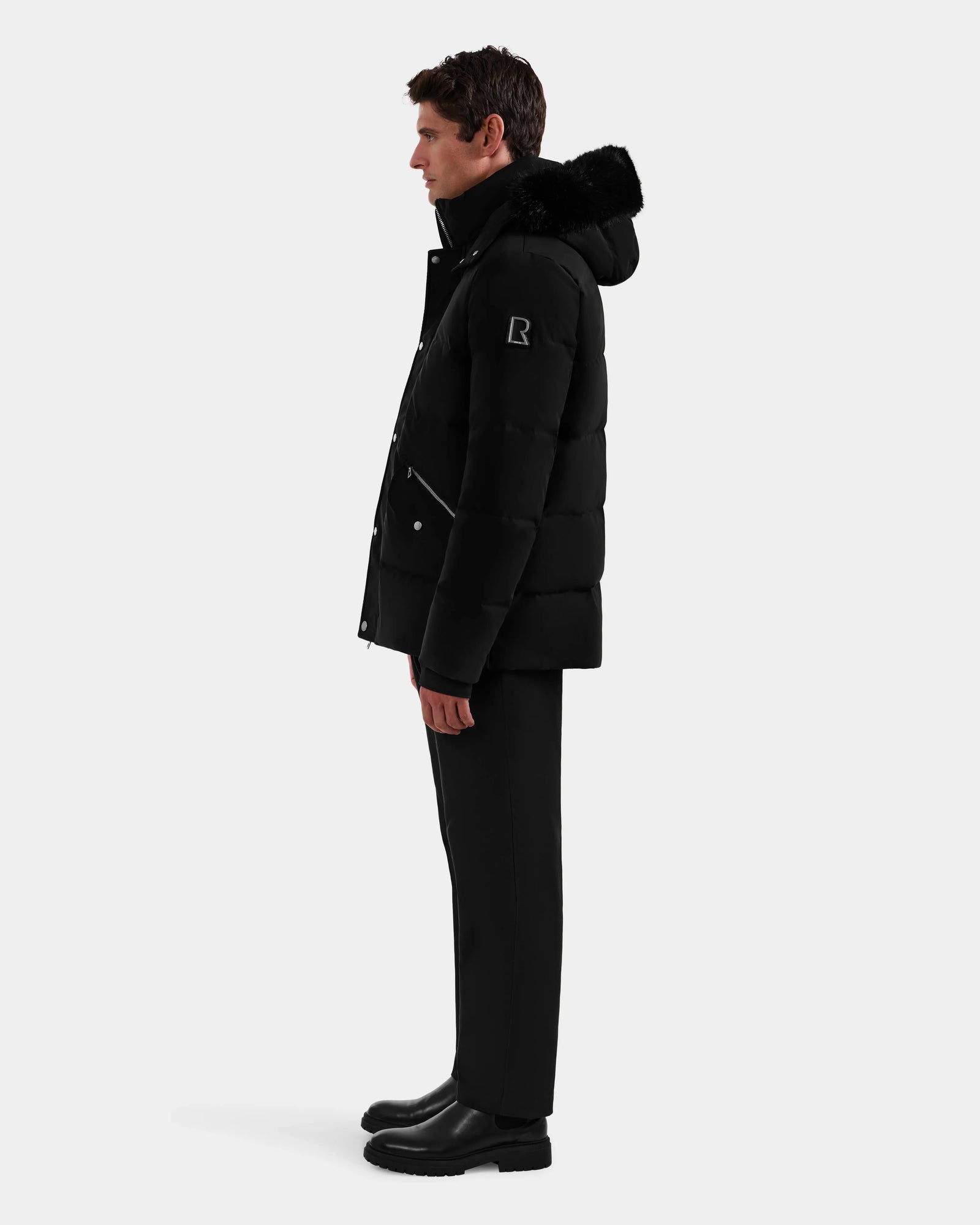 A man stands in profile wearing the Rudsak BRADY - M125510 Black men's quilted down parka with removable hood, paired with black pants and boots, set against a plain light background—a true winter essential.