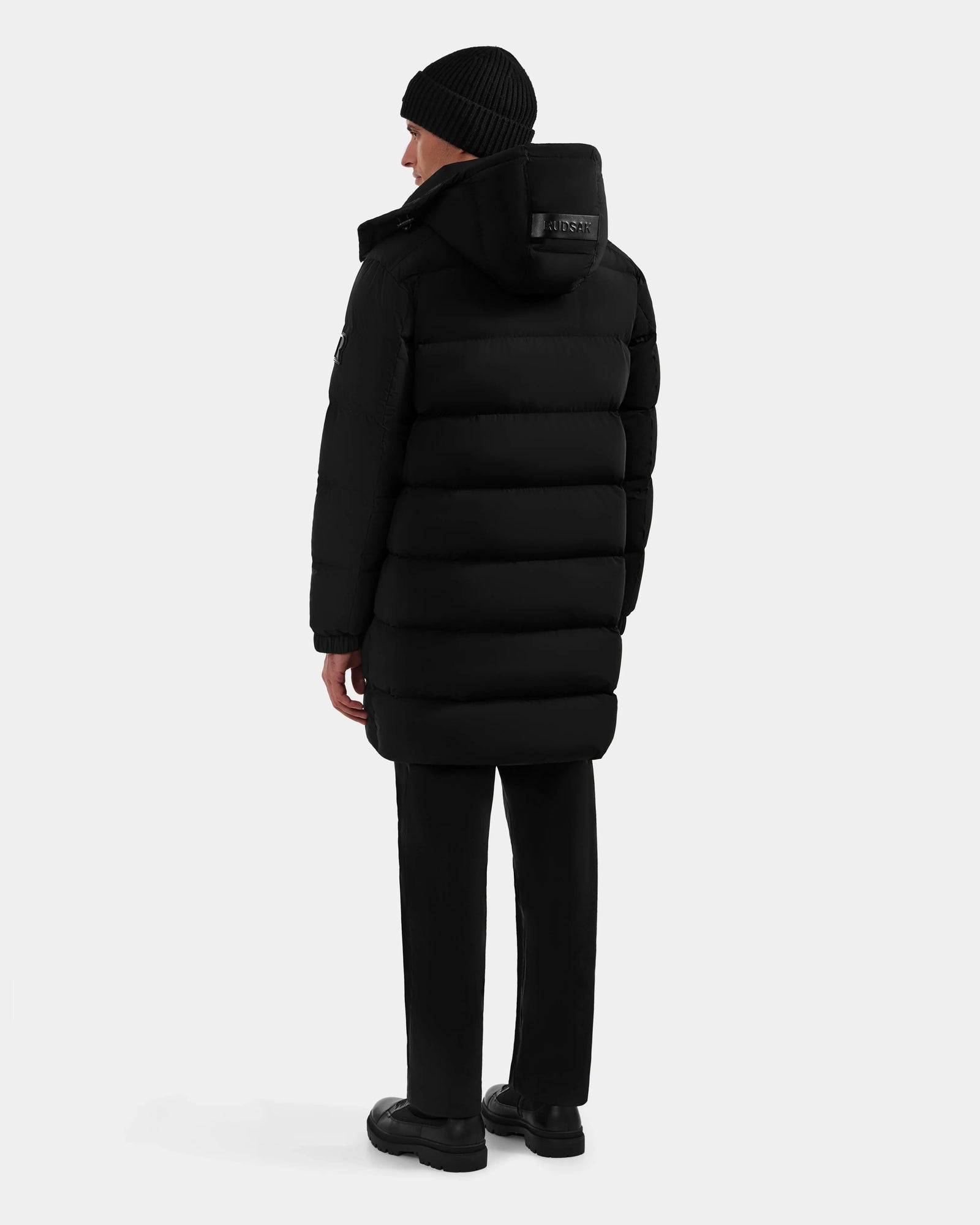 A person wearing the Rudsak BRAYDEN M-M - M125535 Black down puffer coat, black pants, shoes, and a beanie stands facing away against a plain light background, showcasing the coat’s winter versatility.