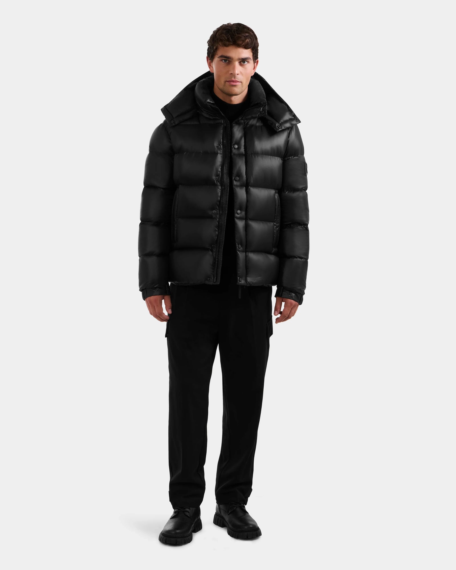 A man faces forward in a shiny black Rudsak BRAYDEN S - M125525 quilted down puffer jacket, paired with black pants and shoes, set against a plain light gray background.