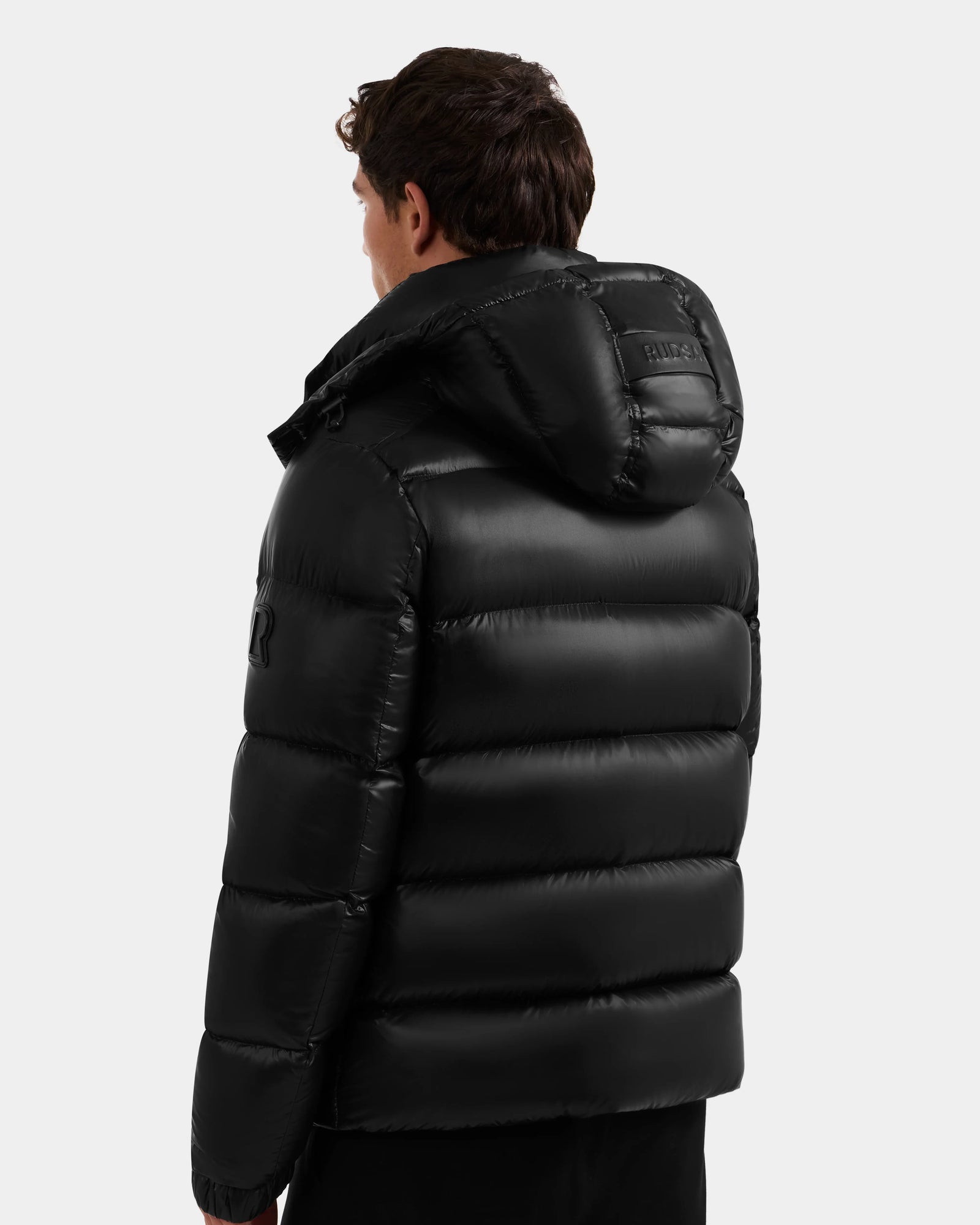 A person with short brown hair is seen from behind wearing the Rudsak BRAYDEN S - M125525 Black men's winter jacket, featuring shiny black quilted down, a hood, horizontal quilting, and a raised logo on the upper arm.