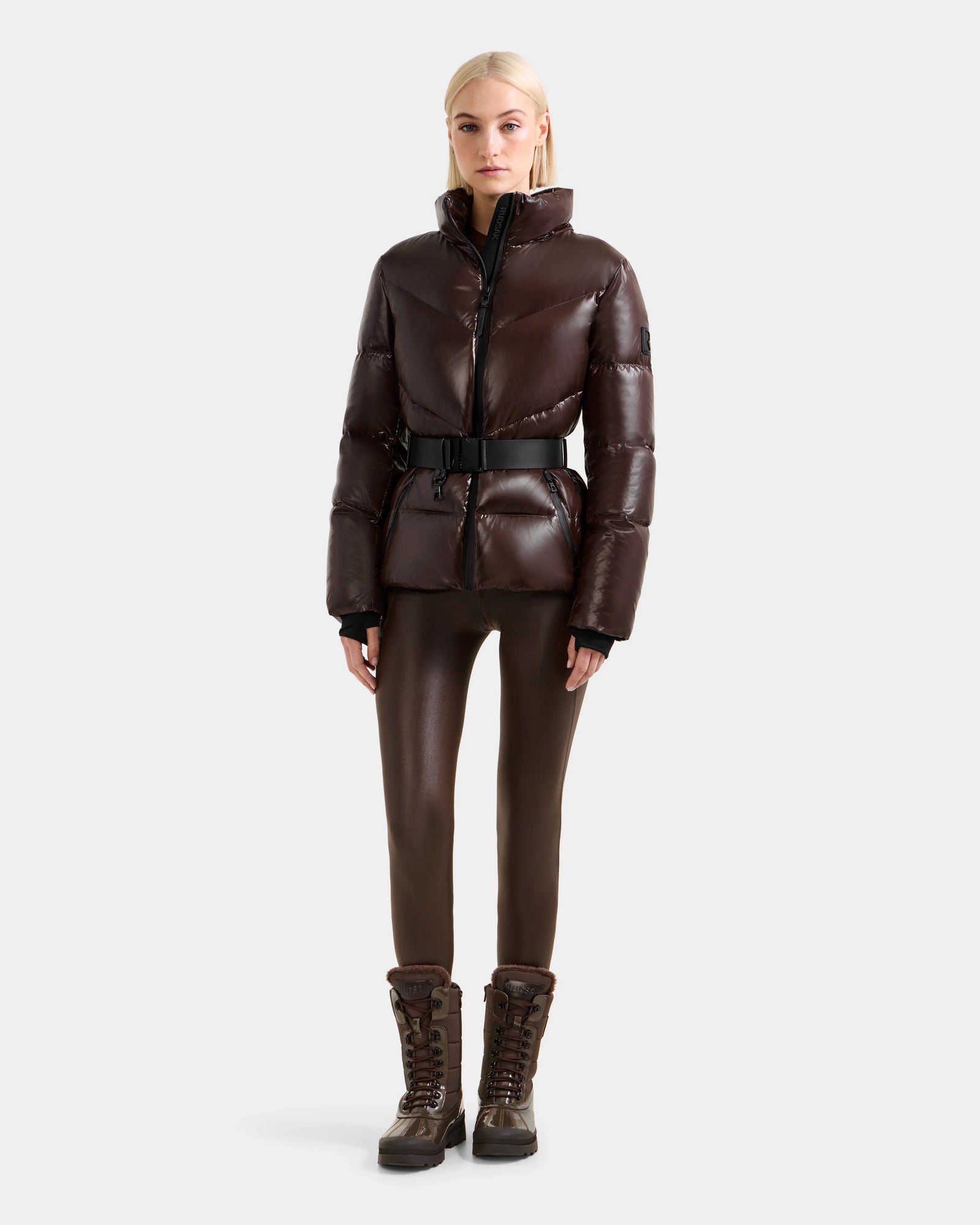 A woman faces forward wearing the Rudsak CHRISTELLE S - L125544 Espresso dark brown down puffer jacket with a belt, fitted pants, and lace-up winter boots against a plain light gray background.