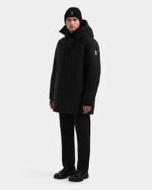 A person wears the Rudsak COREY - M125512 Black men’s down parka and a black beanie, both with a white R logo, plus black pants and shoes. The winter jacket uses RDS-certified down for optimal warmth.