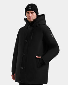 A man stands facing forward against a plain light background, wearing the Rudsak COREY - M125512 Black men’s down parka with a hood and a black beanie, one hand in his pocket.