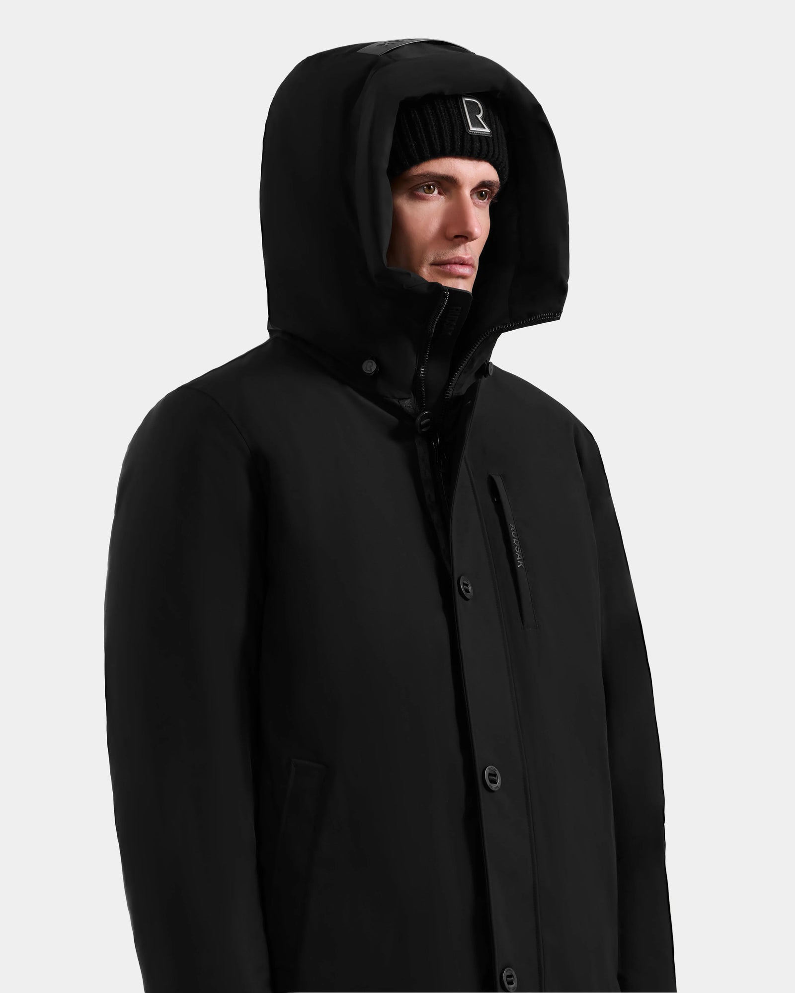 A person wearing the Rudsak COREY - M125512 Black men’s down parka with RDS-certified down and a black beanie featuring a white R logo, looks slightly to the side against a plain light background.