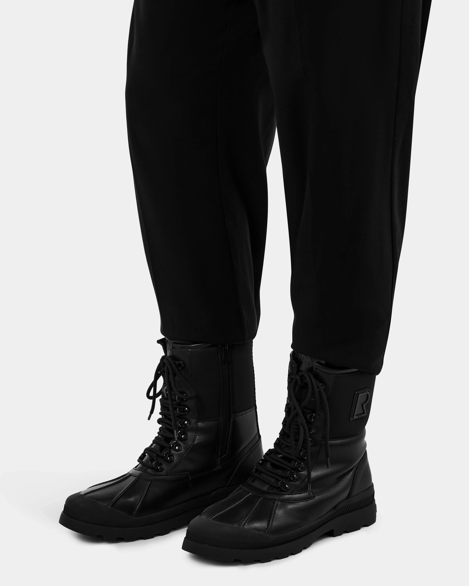 A person stands against a plain white background wearing black pants and Rudsak DAMIEN - M222500 Black men’s winter boots, which feature thick soles and weather-treated leather.