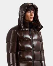A person wearing Rudsak's DANIEL S - M124508 Espresso, a shiny dark brown men's hooded down puffer with a weather-resistant glossy shell and high-loft down fill, hood up and paired with a brown knit beanie, looks to the side against a light background.