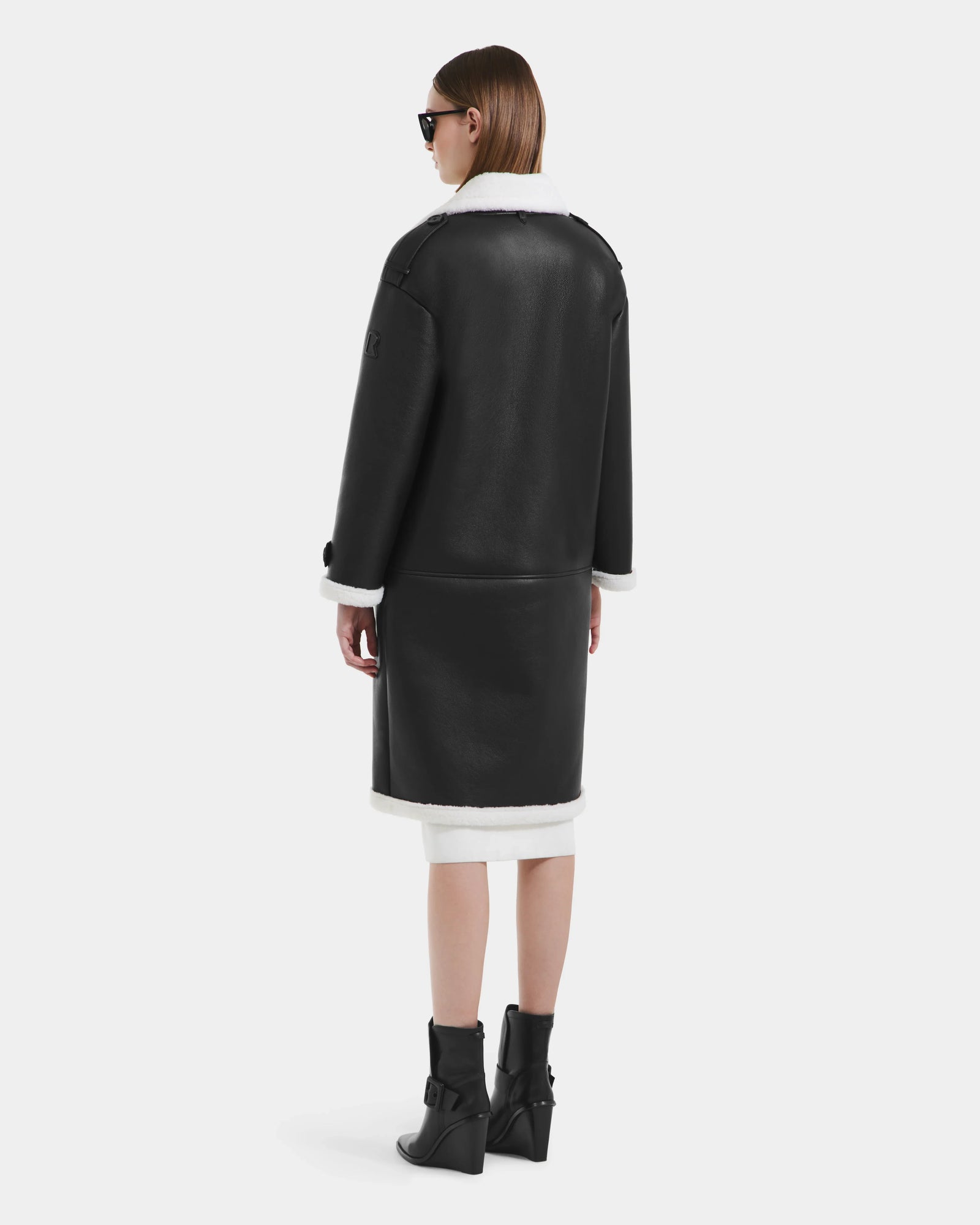 A woman with straight brown hair wears sunglasses and the Rudsak DARCY - L125539 Black vegan leather overcoat with white shearling trim, paired with black ankle boots, standing and facing away from the camera against a plain background.