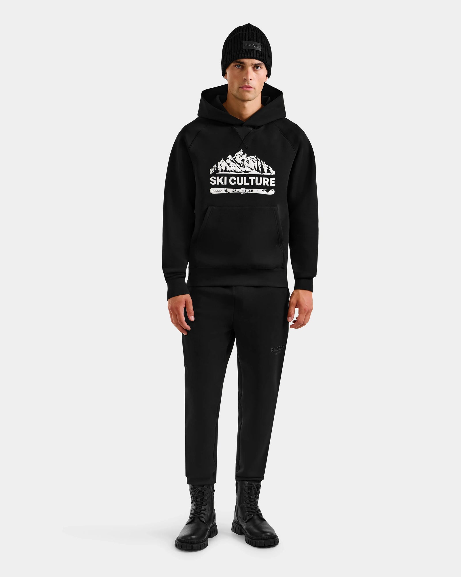 A person stands against a plain background wearing Rudsak’s DARWIN - M525502 Black cotton-blend fleece men’s hoodie with Ski Culture and mountain graphic, paired with a black beanie, black pants, and black boots.