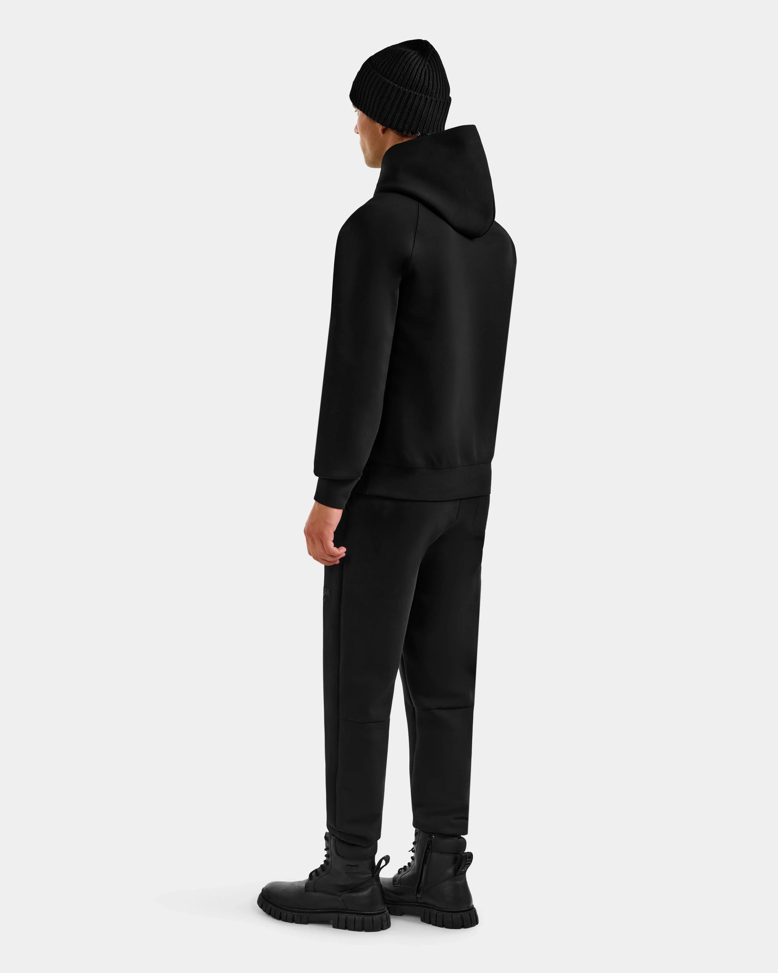 A person stands facing away, wearing the Rudsak DARWIN - M525502 Black men's hoodie with black pants and boots against a plain light gray background.