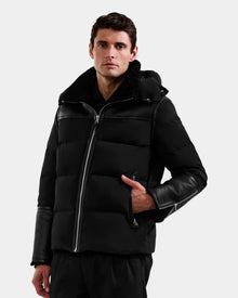 A man wearing the Rudsak DAVID - M125508 Black puffer jacket with leather sleeves and a hood stands against a plain light background, looking directly at the camera with one hand in his pocket.