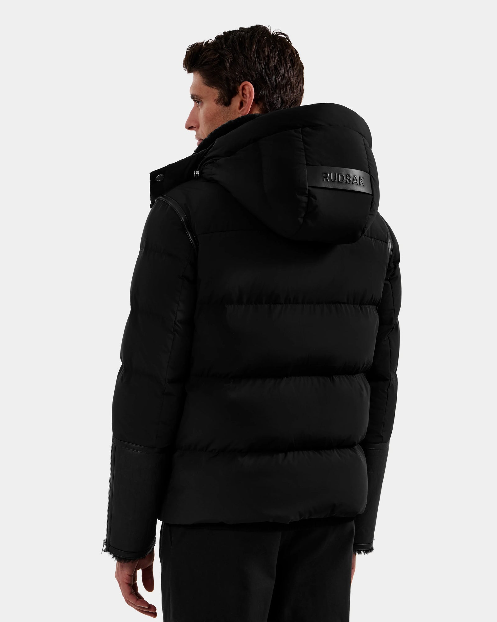 A man with short dark hair wears the Rudsak DAVID - M125508 Black puffer jacket, featuring a visible RUDSAK logo on the back. This men's shearling down puffer is winter-ready, and he faces away from the camera against a plain background.