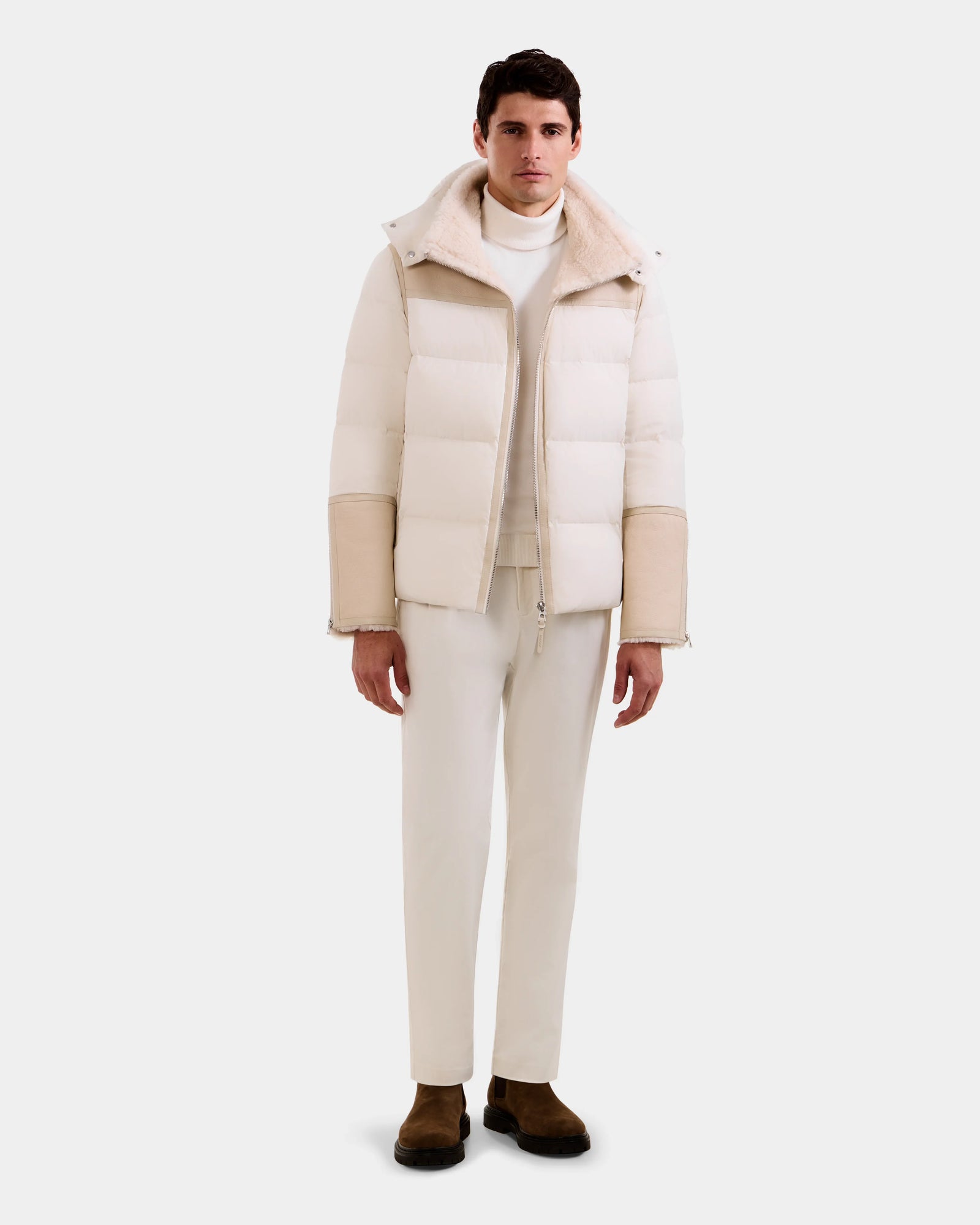 A man stands forward against a white background, wearing the Rudsak DAVID - M125508 Vanilla white down puffer jacket with a beige lamb shearling collar and sleeves, paired with white pants, a turtleneck, and brown suede boots.