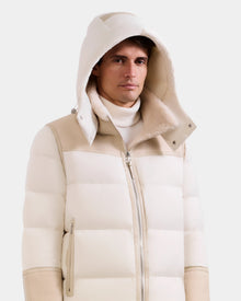 A man wears the Rudsak DAVID - M125508 Vanilla men's shearling puffer jacket with hood and high collar over a white turtleneck, standing against a plain light background.