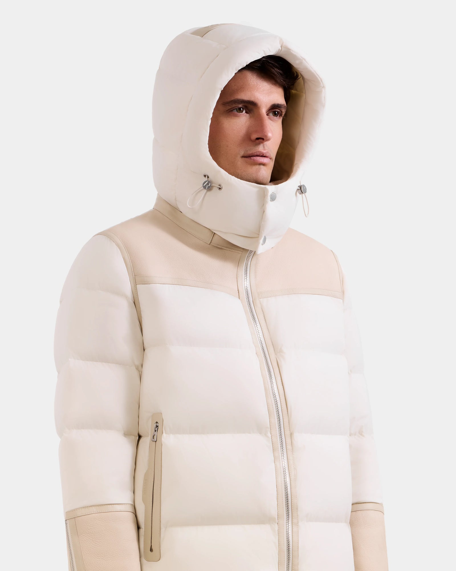 A man wears the Rudsak DAVID - M125508 Vanilla men’s shearling puffer jacket with a hood, high collar, zippered pockets, and drawstring hood, facing slightly right against a plain light background.