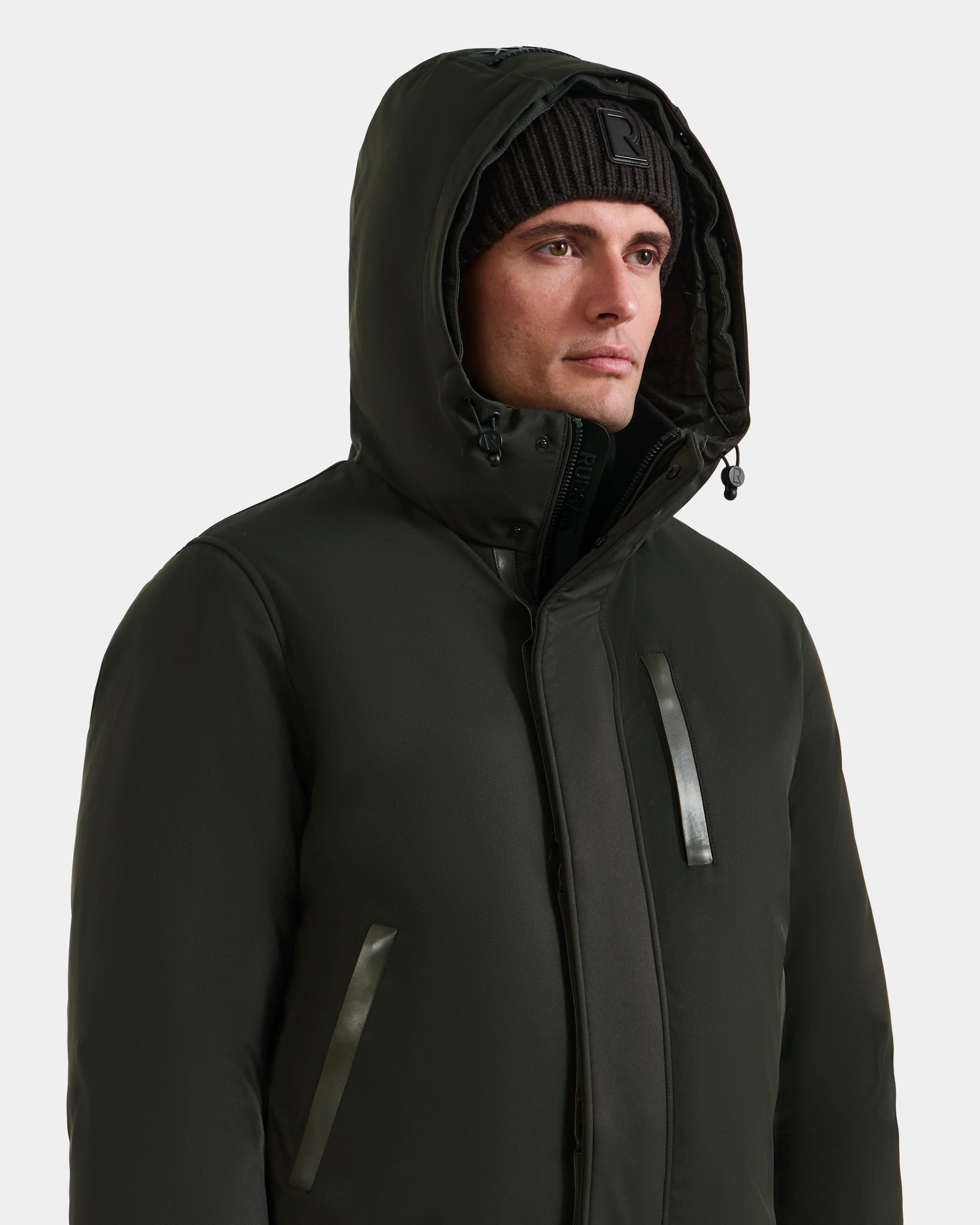 A man wearing the Rudsak DEACON - M123513 Dark Olive bomber-style down puffer jacket and a black beanie looks forward with a neutral expression against a plain light background.
