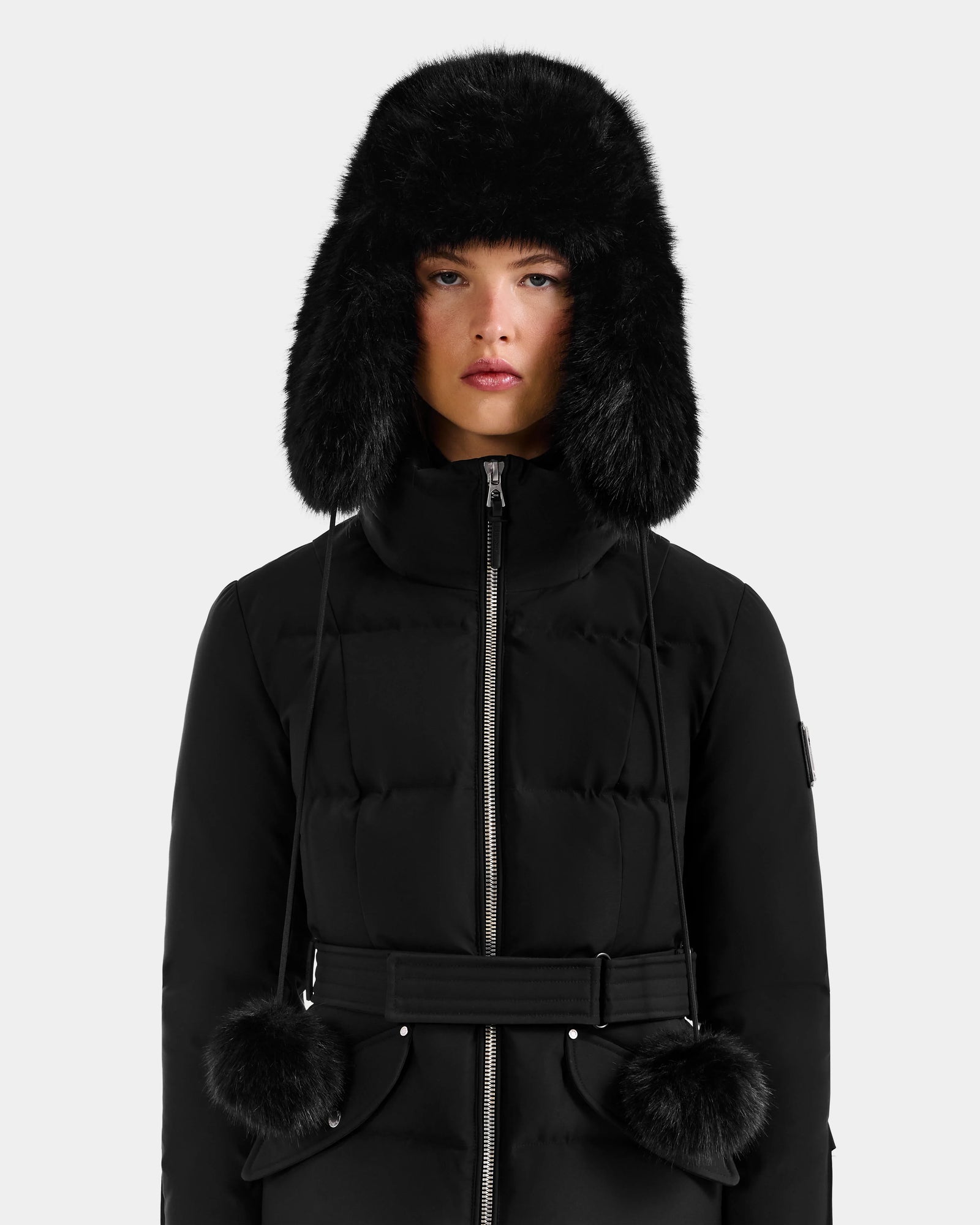 A person wears the Rudsak DEMI - L425539 Black puffer coat with a belted waist and a Rudsak faux-fur aviator hat with ear flaps and pom-pom ties, standing against a plain light gray background.