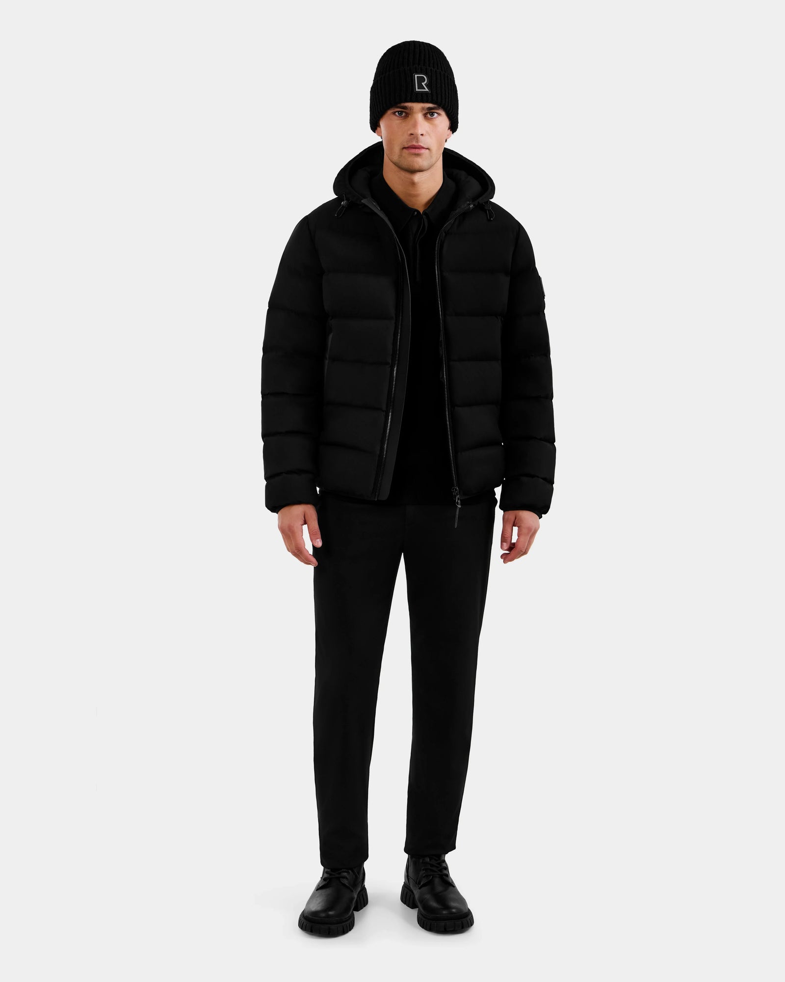 A man stands facing forward, wearing the Rudsak DEMIAN M - M123507 Black down puffer, paired with a black beanie, black pants, and black shoes against a plain white background.