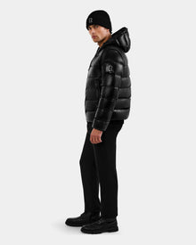A man in the Rudsak DEMIAN S-R - M123505 Black down puffer jacket, black beanie, pants, and boots stands sideways against a plain light background, looking at the camera.