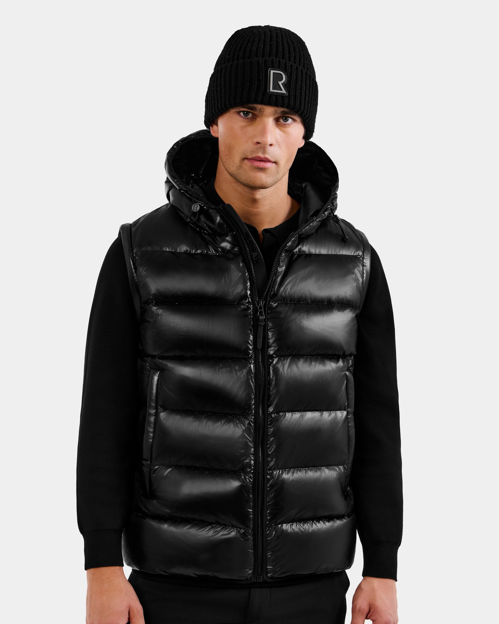 A man in a Rudsak DEMIAN S-R - M123505 Black 2-in-1 puffer jacket with hood, black knit beanie, and black long-sleeve shirt stands facing forward against a plain light gray background.