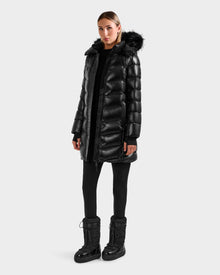 A woman stands against a plain background wearing the Rudsak ELISA S - L125572 Black quilted puffer jacket with a fur-trimmed hood, paired with black leggings and winter boots, looking straight ahead with a neutral expression.